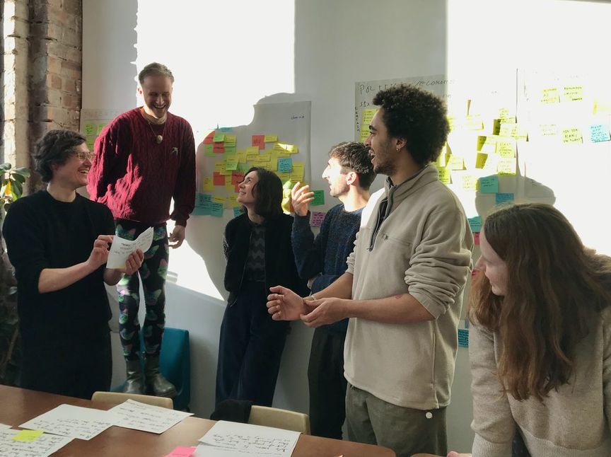 A photo of the team from the Glasgow retreat, they are in conversation around a wall of sticky notes, it is sunny and everyone is smiling.
